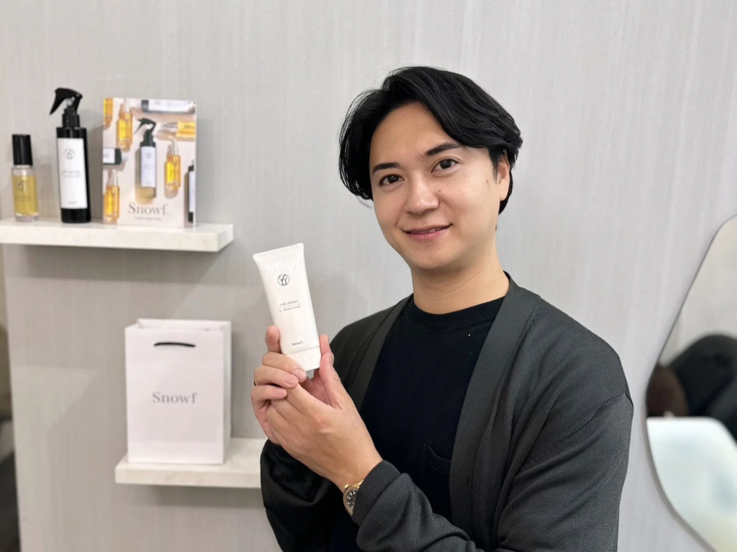 若い男性が笑顔でカメラを見つめ、白いチューブ状の美容製品を手に持っています。