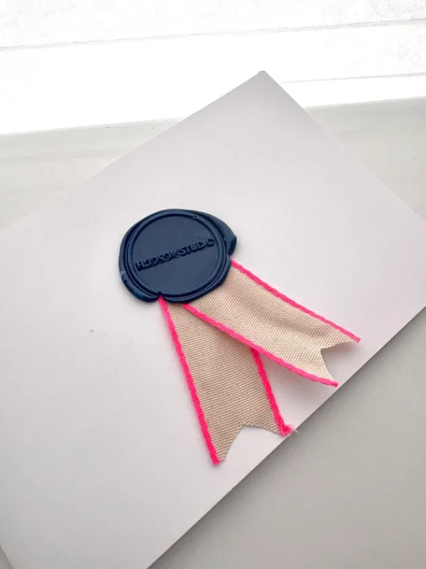 白いカードにネイビーのシーリングワックスとベージュとネオンピンクのリボンが飾られています。シーリングワックスには「HUDSON STUDIO」の文字が刻まれています。