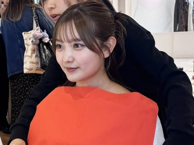 黒い服を着た若い女性が、オレンジ色の布を首元に当て、髪をポニーテールにまとめている