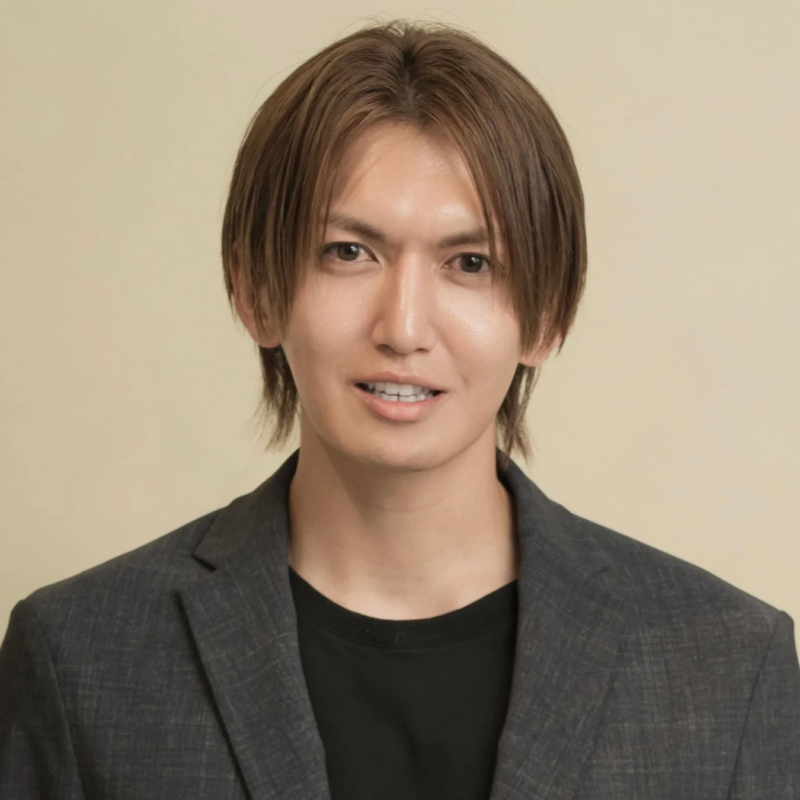 茶色の髪とグレーのジャケットを着用した若い男性が、カメラに向かって微笑んでいるポートレート写真