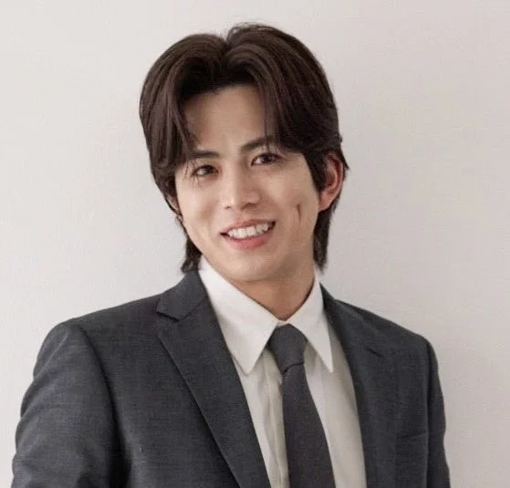 ダークスーツとネクタイを着用した若い男性が、白い背景の前でカメラに向かって笑顔を見せているポートレート