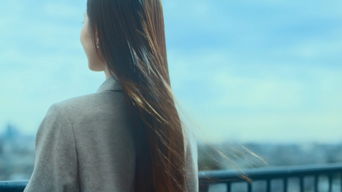 風になびく長い髪の女性が、青空と遠くの街並みを背景に、後ろ姿で佇んでいます