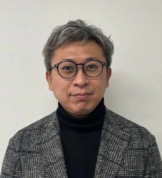 眼鏡をかけた白髪の日本人男性が、黒いタートルネックとグレーのチェック柄ジャケットを着用し、正面を向いて立っている上半身のポートレート写真です。