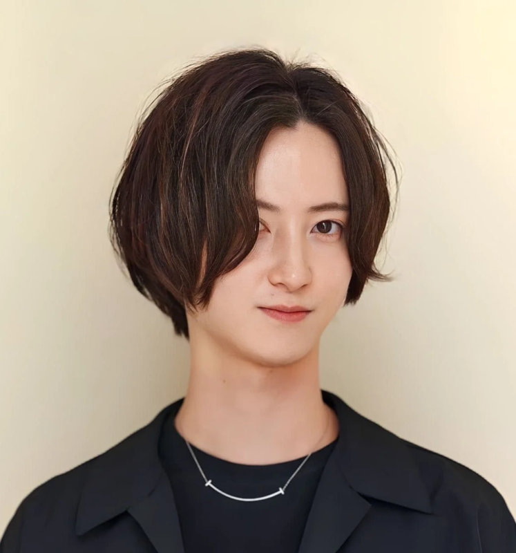 明るい背景の前で、黒いシャツとジャケットを着用し、スマイルマークのネックレスをつけた男性のポートレート