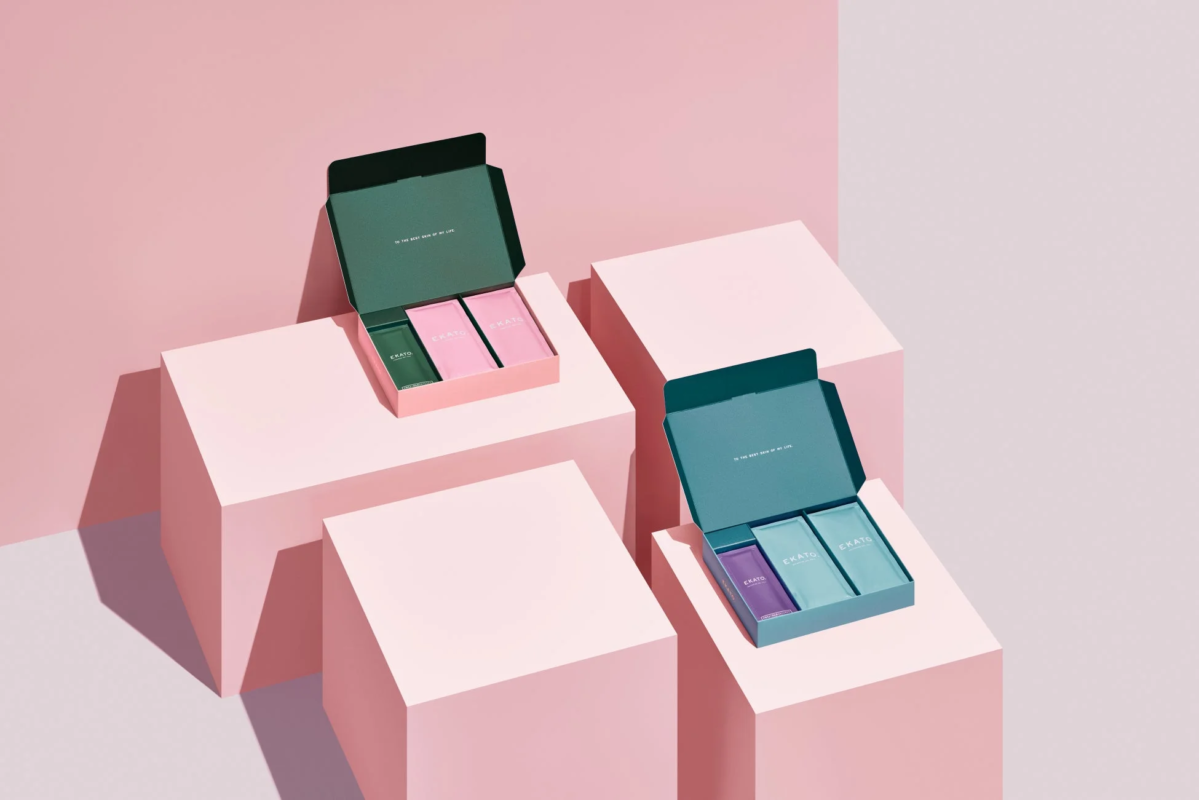 ピンクとティールグリーンの2つの開いたギフトボックスが、ピンクの幾何学的な台座に置かれています。各ボックスには「EKATO」と書かれた3つの小さなパッケージが入っており、蓋には「TO THE BEST SKIN OF MY LIFE.」と書かれています。