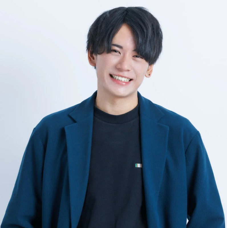 白い背景の前で、青いジャケットと黒いTシャツを着た若い男性が、明るい笑顔を見せています。