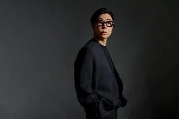 黒い服とメガネをかけた男性が、暗い背景を背にこちらを見つめているポートレート写真