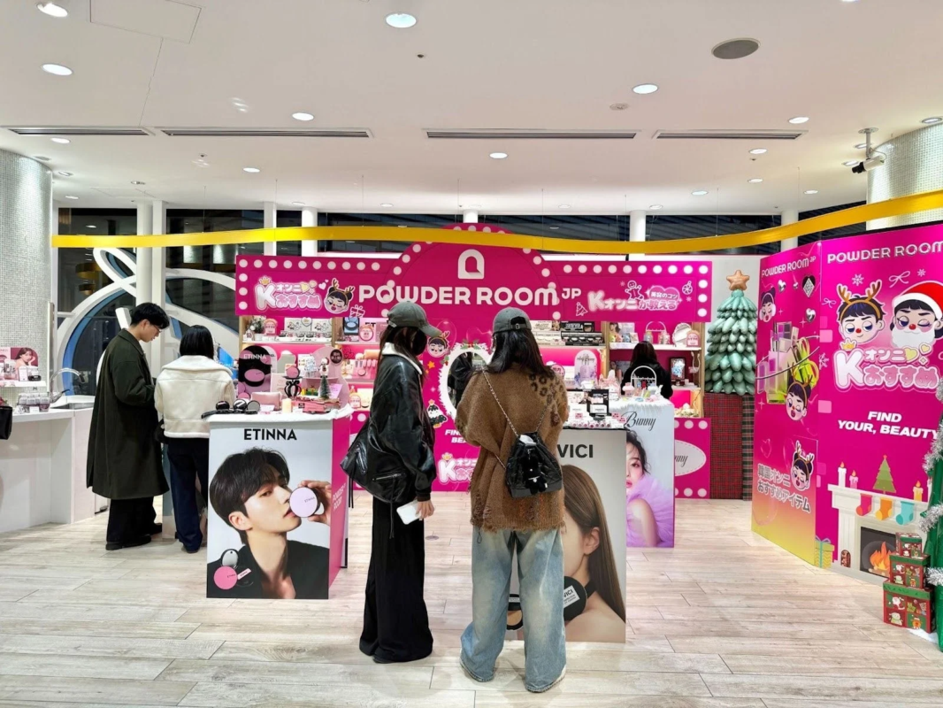 ピンクを基調とした「POWDER ROOM JP」のポップアップストアで、人々がコスメやビューティー製品を熱心に見て回っています。クリスマス装飾が施され、K-オンニおすすめの韓国コスメが多数展示されており、賑やかなイベントスペースの様子が伺えます。