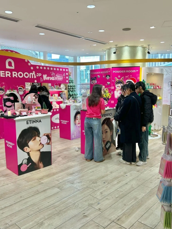 商業施設内で開催されている、ピンクがテーマのコスメポップアップストアの様子。ETINNAやVIDIVICIなどのブランドが並び、クリスマスツリーも設置され、数人の若者が商品を見て賑わっています。