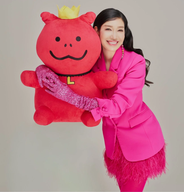 鮮やかなピンクのブレザーとスパンコールの手袋を身につけた女性が、王冠と「L」のマークが付いた赤い大きなぬいぐるみを抱きしめ、笑顔を見せている。