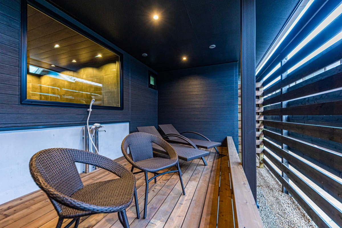 モダンなデザインの屋外テラス。木製デッキにはリラックスできるラウンジチェアや椅子が並び、落ち着いた雰囲気です。窓に映る屋内とシャワーから、サウナやプライベートバスに隣接する宿泊施設の一部と推測されます。