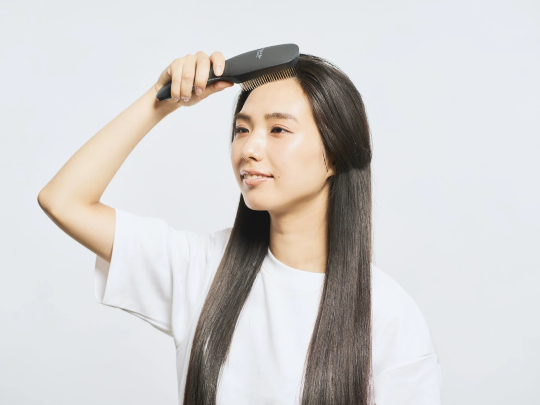 白いTシャツを着たアジア人女性が、黒いブラシで長くまっすぐな髪をとかしている様子。