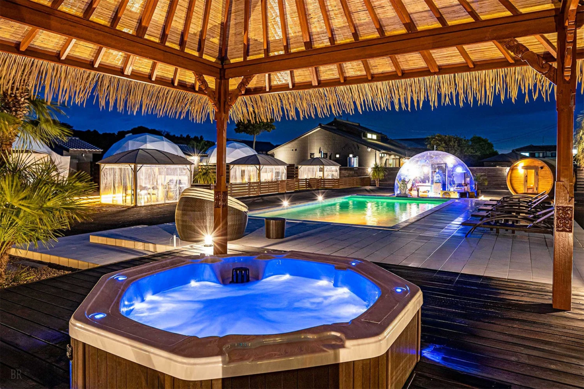 夜の豪華なリゾート施設で、青く光るジャグジー、カラフルにライトアップされたプール、複数のグランピングドーム、そして樽型サウナが配置されています。リラックスできる休暇の雰囲気が漂っています。