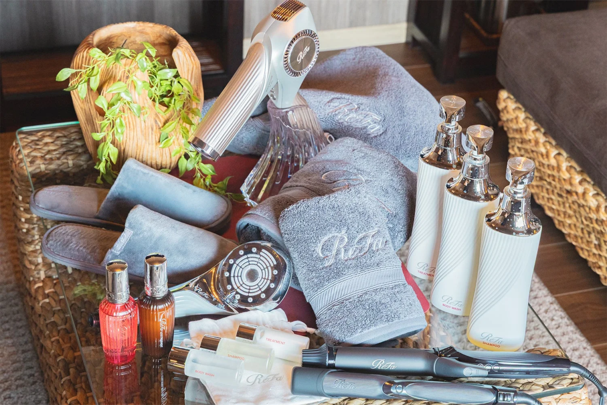 Refaブランドの美容家電(ヘアドライヤー、ヘアアイロン、シャワーヘッド)や、ロゴ入りタオル、スリッパ、シャンプー、ボディウォッシュ、トリートメントなどのアメニティが並べられた画像です。ホテルやスパの客室に用意された上質なアイテム群を示しています。