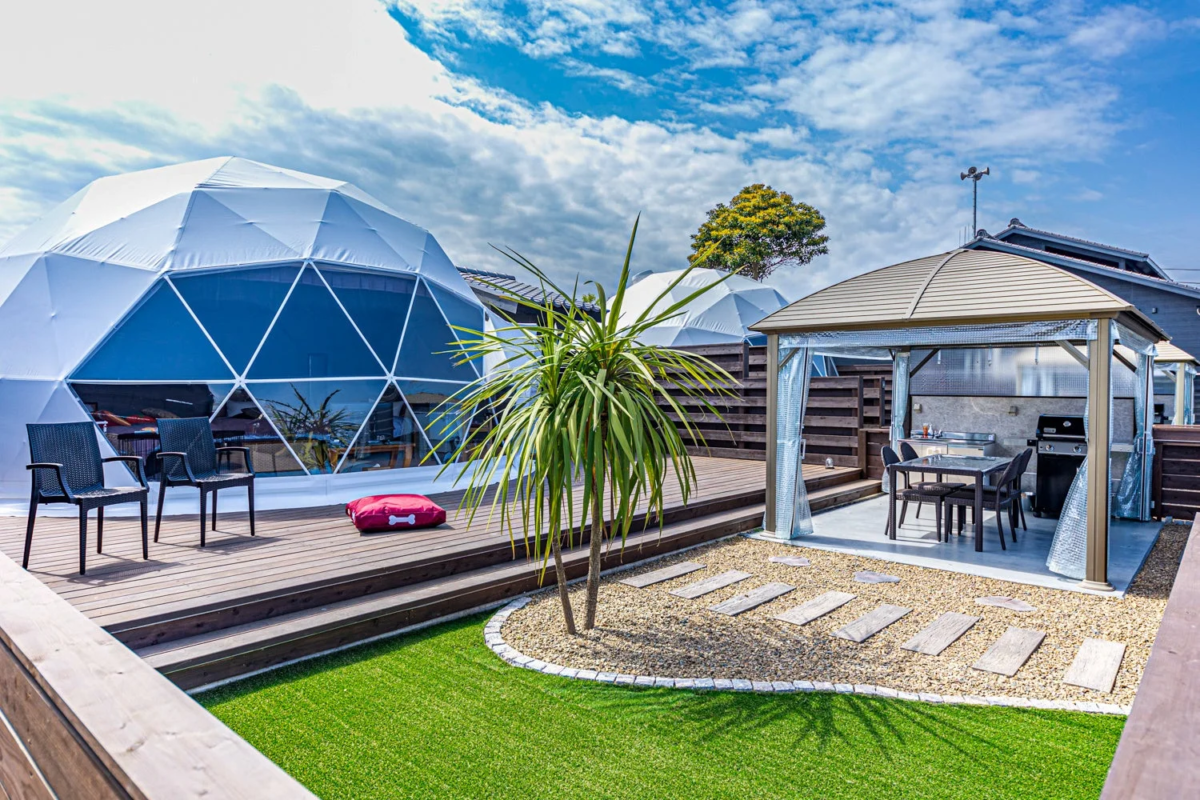 白いドーム型テントとバーベキュー設備のある屋外スペースが広がる、グランピング施設のような風景です。晴れた日の開放的な雰囲気が伝わります。