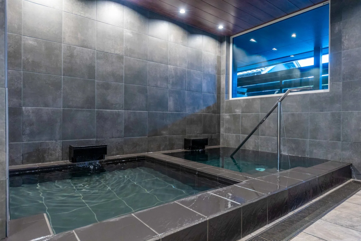ダークグレーのタイル張りの壁と木目調の天井が特徴のモダンな内湯です。2つの浴槽があり、窓からは外の景色が望めます。手すりも設置されており、リラックスできる空間です。