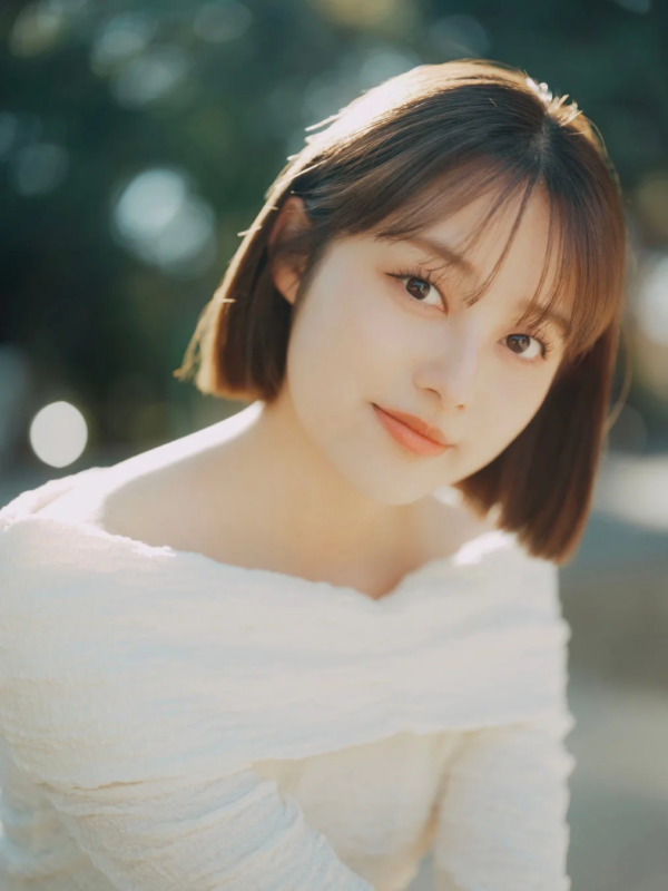 明るい自然光の下、白いオフショルダーの服を着たショートヘアの女性が、穏やかな表情でカメラを見つめているポートレート写真です。背景は柔らかくボケています。