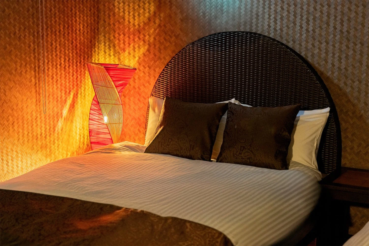 温かい間接照明が心地よい寝室の様子です。ベッドには濃色のクッションが置かれ、特徴的な赤いランプが柔らかな光を放ち、壁の編み込みテクスチャがリラックスできるアジアンテイストの空間を演出しています。