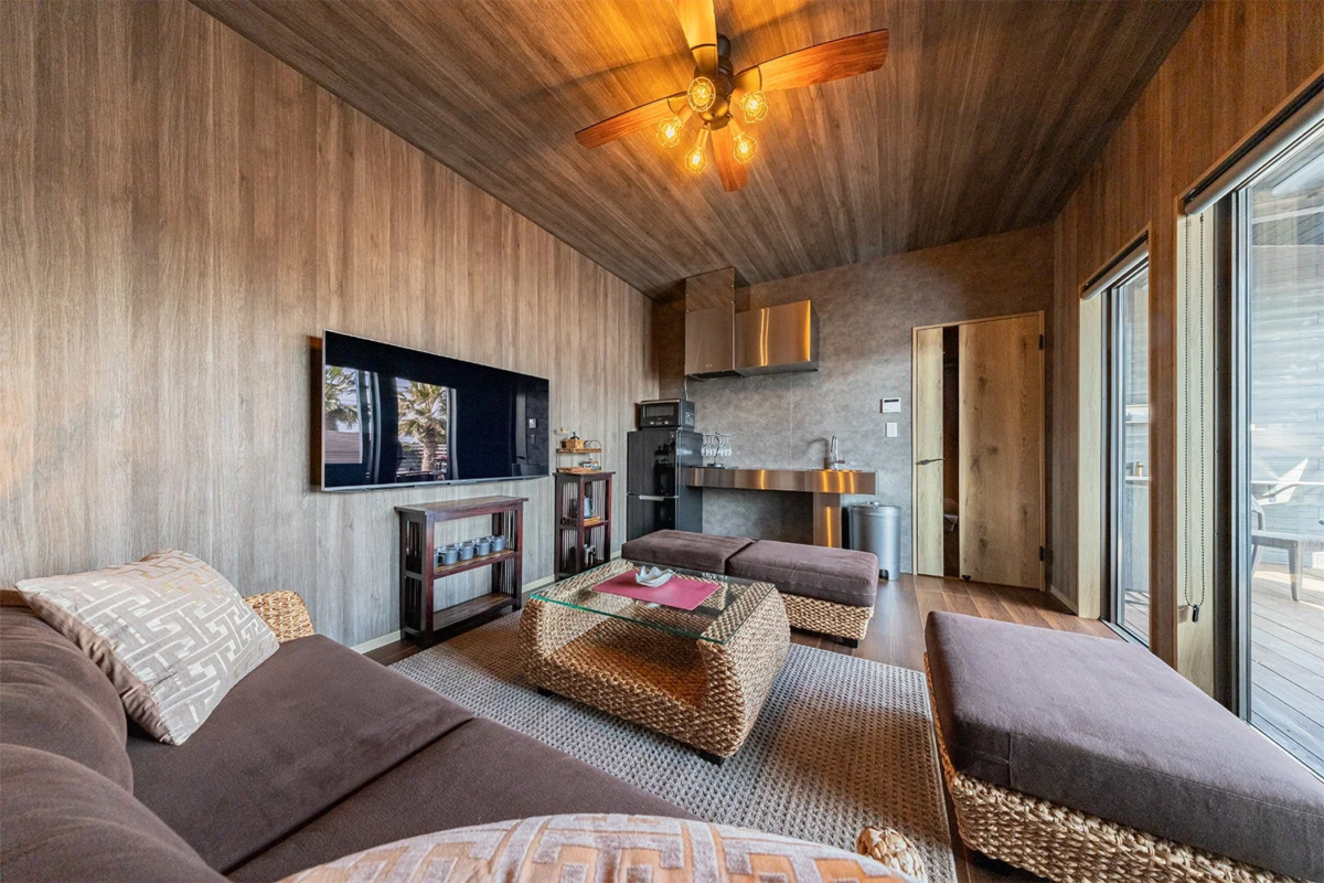 木目調の壁と天井が特徴的なモダンなリビング兼キッチン空間です。シーリングファン、大型テレビ、ソファ、そして屋外に続く大きな窓があり、リラックスできるリゾートのような雰囲気が漂っています。