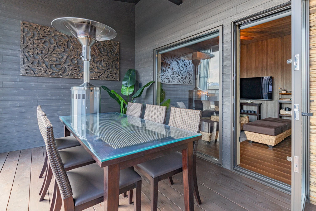 モダンな屋外テラスの様子で、ガラス天板のダイニングテーブルと椅子、パティオヒーターが設置されています。壁には装飾アートがあり、隣接する室内の一部も開いたドアから見えます。
