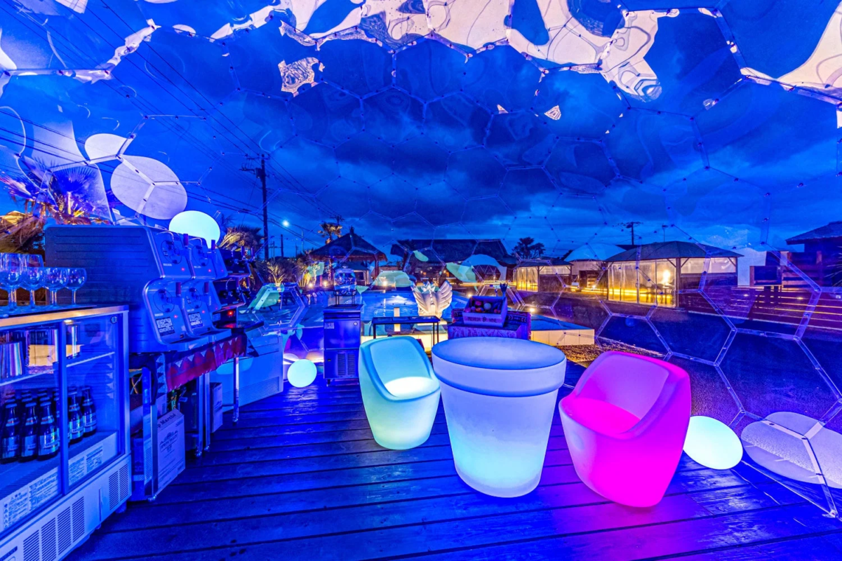青い光に包まれた未来的な雰囲気の屋外ナイトラウンジ。六角形のドーム構造の下、光る椅子やバーカウンターが配置され、幻想的な空間を演出しています。