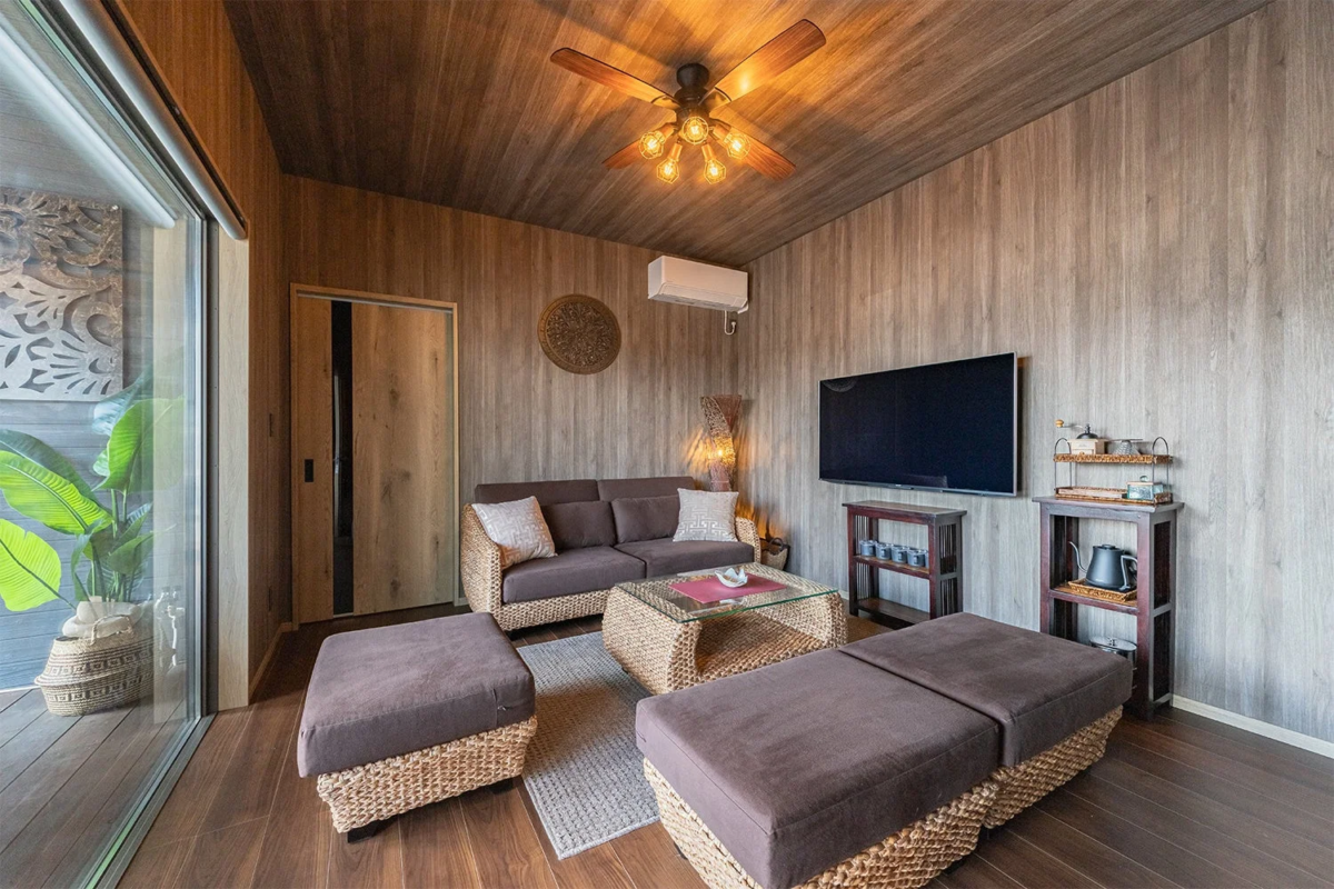 木目調の壁と天井が特徴的な、リゾート感あふれるリビングルームです。ラタン素材のソファやオットマン、ガラス天板のコーヒーテーブルが配置され、シーリングファンや観葉植物がトロピカルな雰囲気を演出しています。大型テレビや間接照明も備え、快適で落ち着いた空間です。