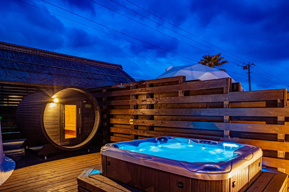 夜の屋外に設置されたバレルサウナと、青く光るジャグジーが特徴的な空間です。木製デッキの上でリラックスできるプライベートなひとときを演出しており、落ち着いた雰囲気の中でくつろぎの時間を過ごせそうです。