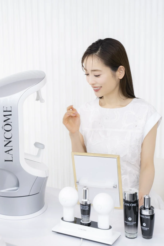 白い背景の部屋で、笑顔の女性がランコムの美容機器と化粧品に囲まれています