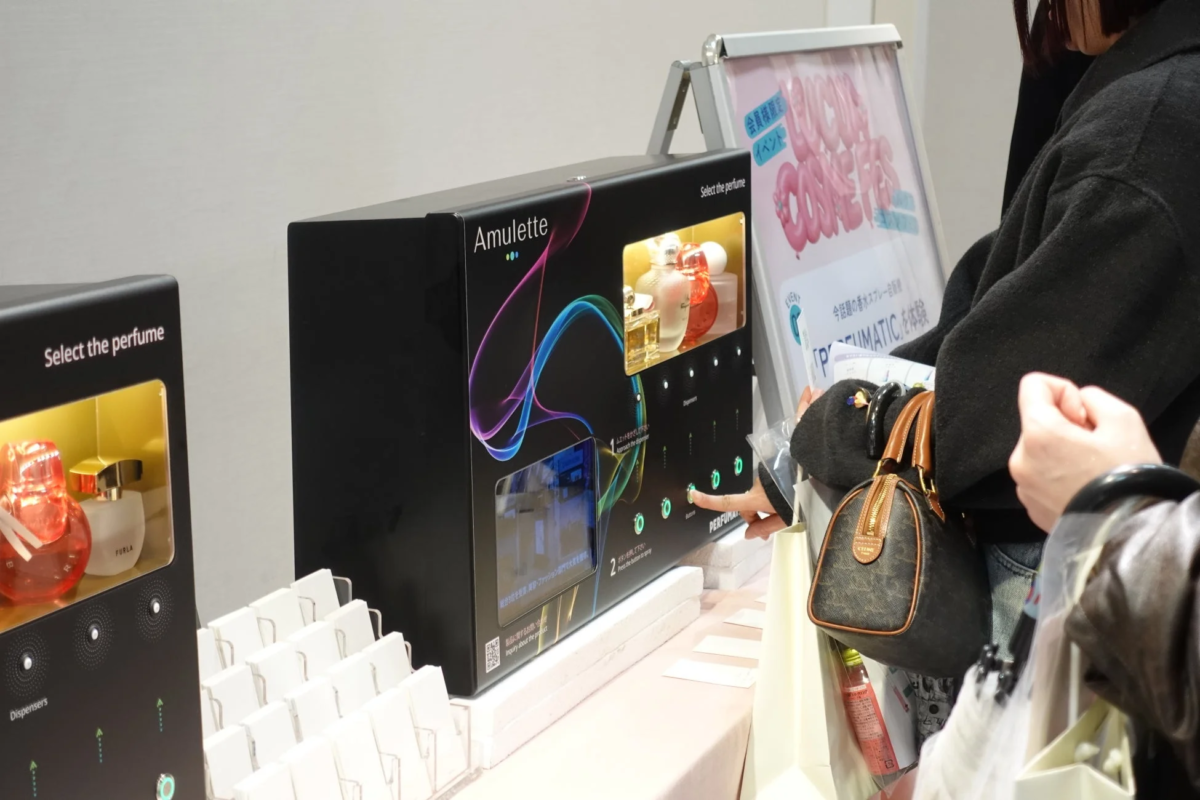 店舗で、女性が「Amulette」という自動香水ディスペンサーを操作している様子。FURLAなどの香水を試せる体験イベントが開催されており、製品情報も提供されています。