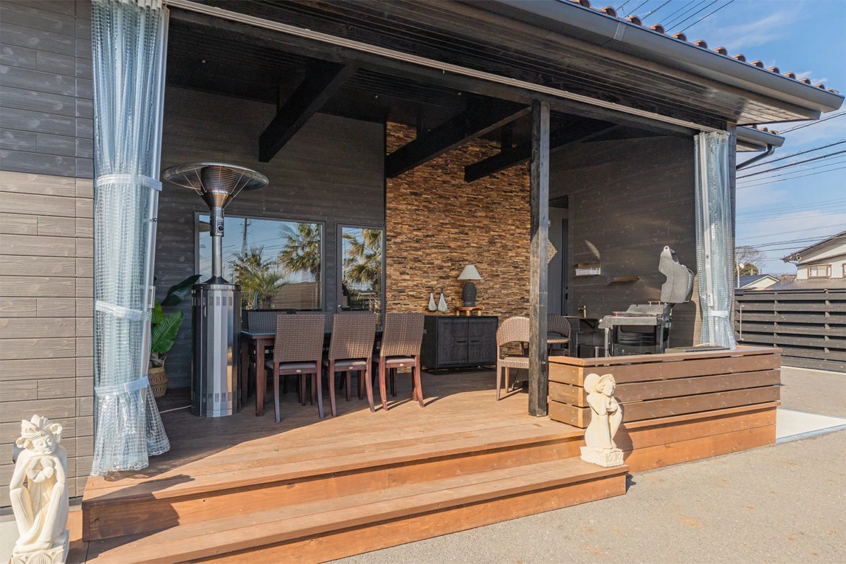 晴れた日の屋外リビング空間。ウッドデッキにはダイニングセットと屋外ヒーターが配置され、石壁やリゾート風の装飾が特徴的な、くつろぎの場です。