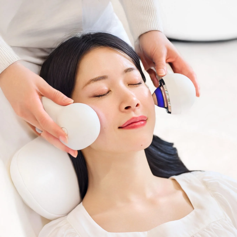 女性が目を閉じてリラックスした状態で、両手で持った美顔器を使ったフェイシャルエステを受けている様子