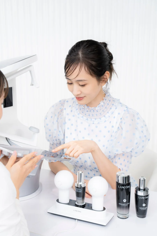 水玉模様のブラウスを着た女性が、ビューティーカウンセラーからタブレットを使ってスキンケアに関する説明を受けています