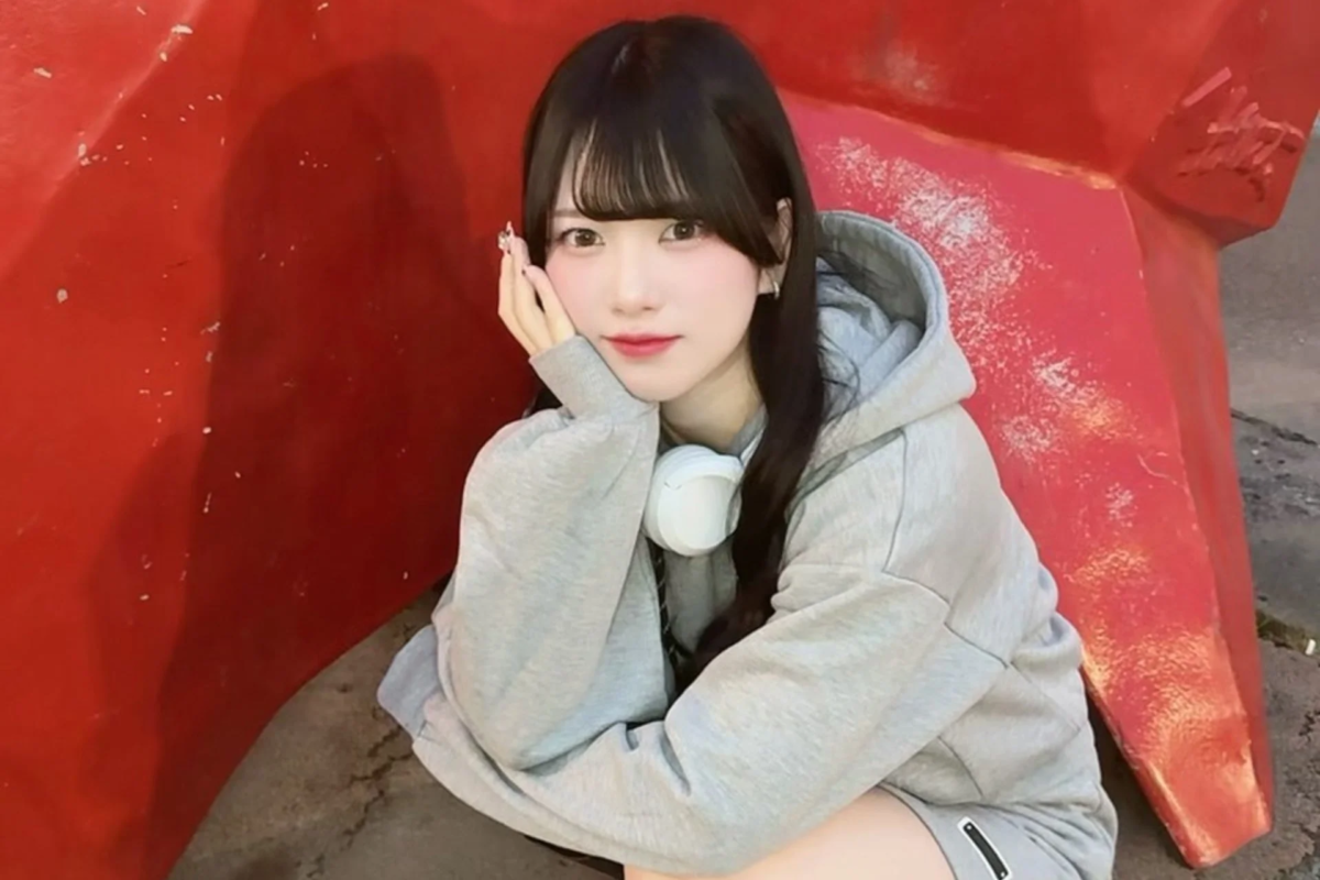 赤い背景の前で、グレーのパーカーを着てヘッドホンを首にかけた若い女性が、頬に手を当ててカメラを見つめているポートレート写真です。カジュアルで可愛らしい雰囲気が特徴です。