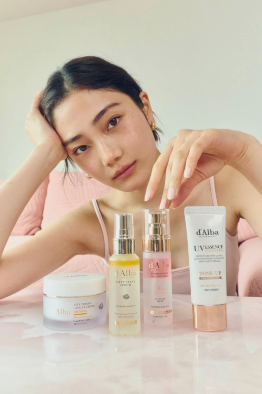 d'Albaのスキンケア製品がテーブルに並べられ、若い女性がリラックスした表情でカメラを見ている