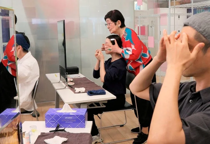 美容教室で、高齢の女性が若い女性にフェイシャルケアやメイクアップの指導をしている様子