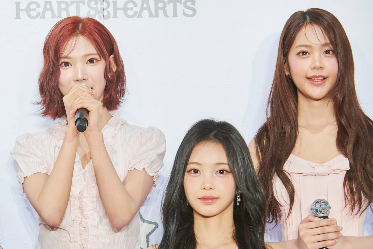 白い背景の前に3人の若い女性アイドルが立っており、2人がマイクを持っています