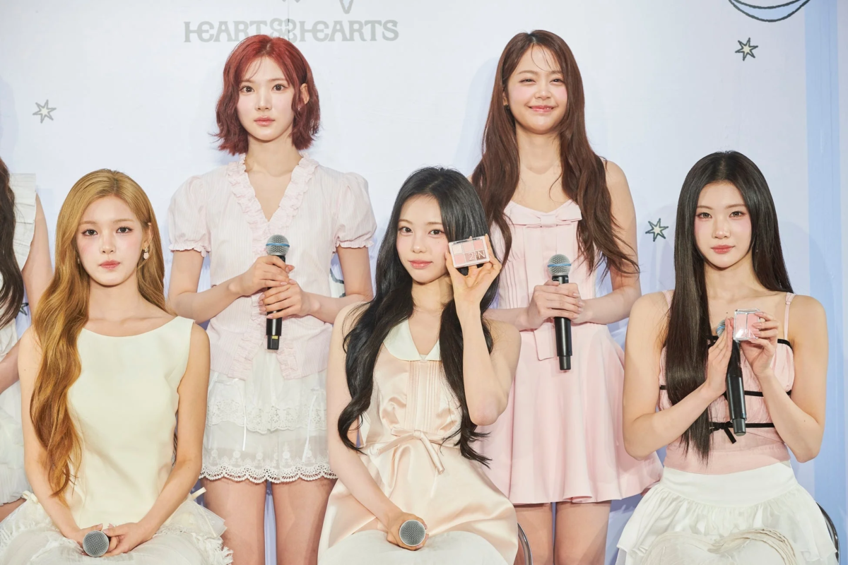 5人の女性アイドルグループが、イベントでマイクを持ち、笑顔で化粧品をプロモーションしている様子を捉えた集合写真