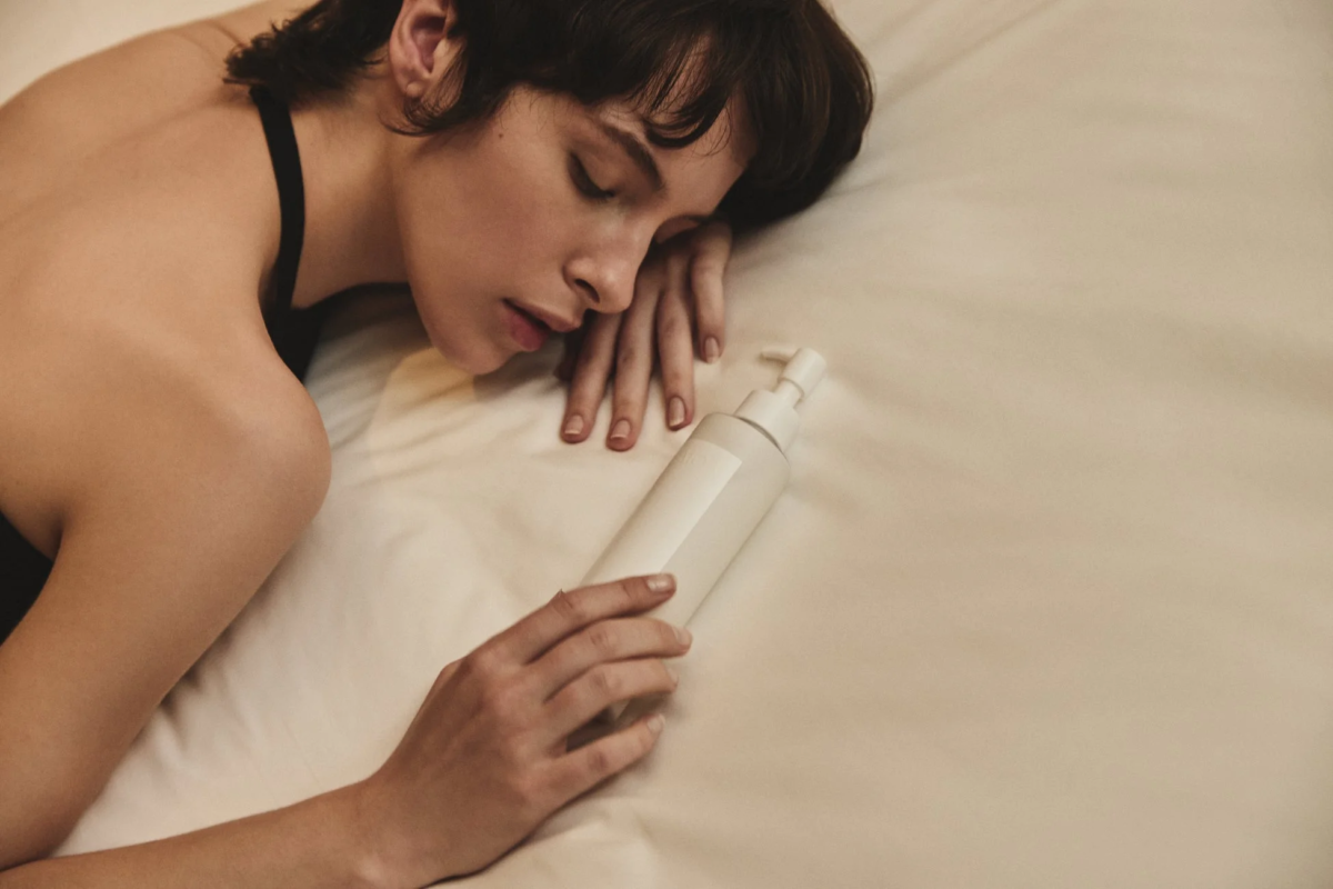 ベッドに横たわり目を閉じた女性が、白いポンプ式ボトルに手を添えています。