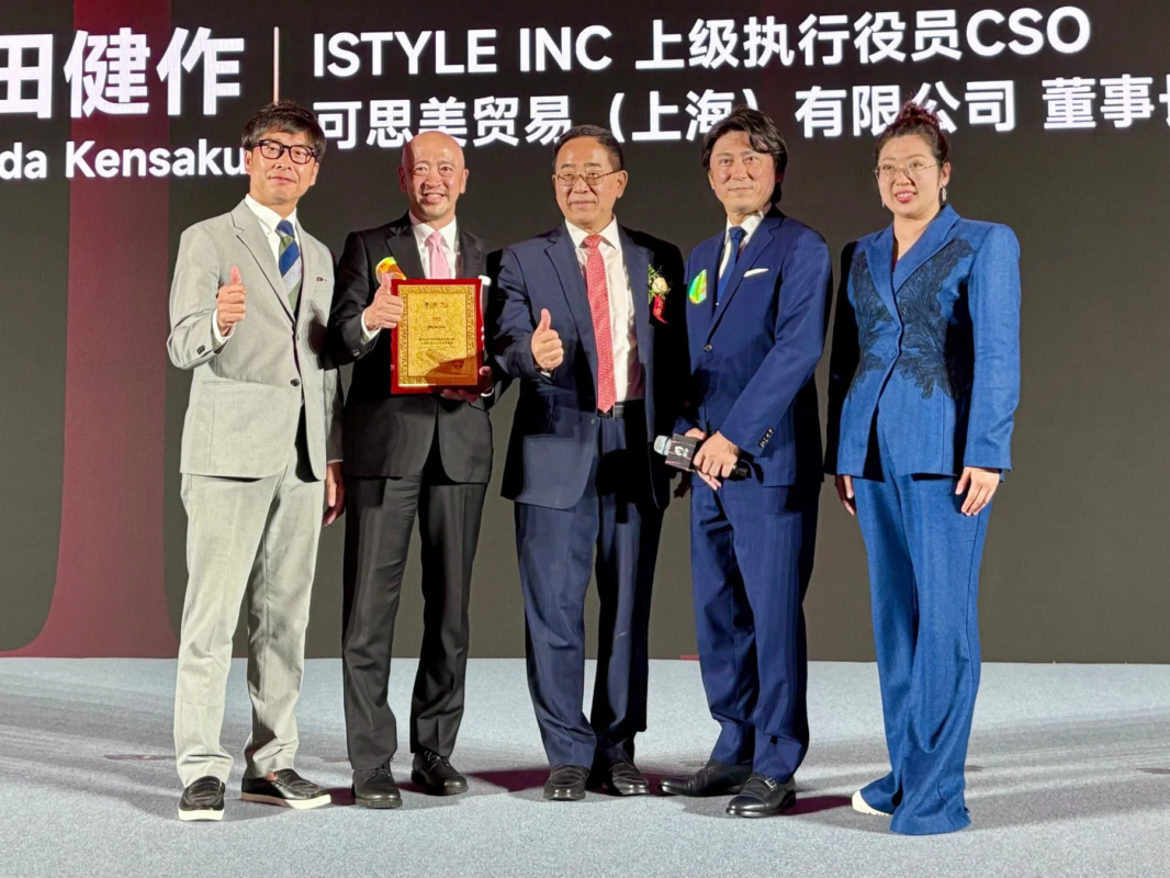 ビジネスイベントのステージで、5人の男女が並んでポーズをとっています。中央の男性は賞状を持ち、他の数名も親指を立てており、表彰式か記念撮影のようです。背景には企業のロゴと役職名が書かれています。