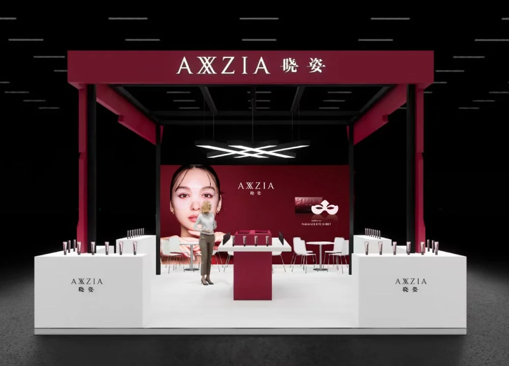 コスメブランドAXXZIA(暁姿)のモダンな展示ブース。大きな女性の顔のディスプレイと、アイシートなどの製品が並べられている。赤と白を基調としたスタイリッシュなデザインが特徴。