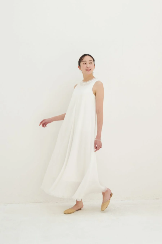 白いノースリーブのマキシ丈ワンピースを着た女性が、白い背景の前で優しく微笑みながら歩いているポートレート。足元は麦わら素材のフラットシューズで、全体的にクリーンでリラックスした印象です。