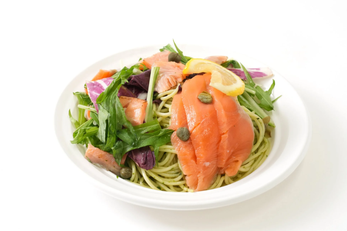緑色のパスタにスモークサーモン、新鮮な葉物野菜、ケッパー、レモンスライスが盛り付けられた冷製パスタサラダの画像です。白い皿に盛られており、健康的で彩り豊かな一皿が食欲をそそります。