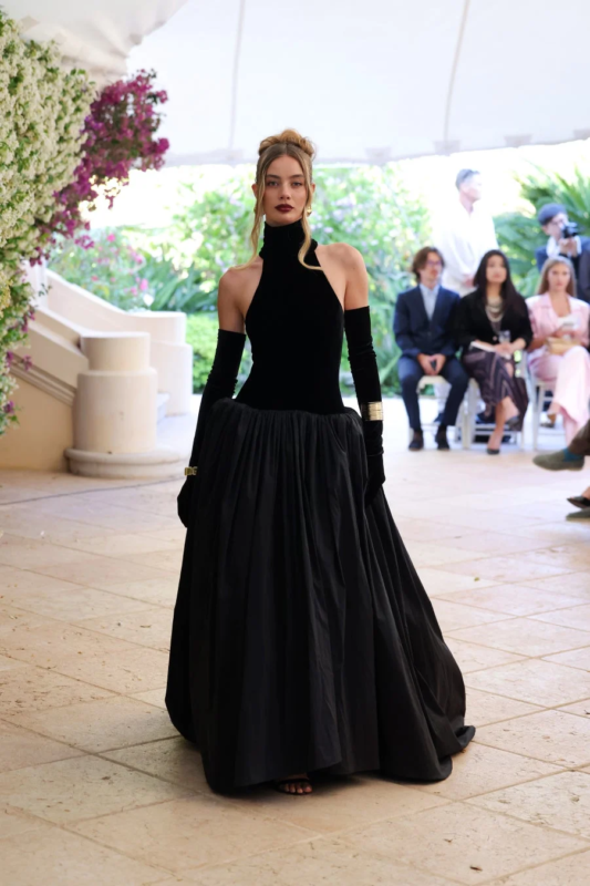 黒いホルターネックのロングドレスと長い手袋を身につけたモデルが、屋外のファッションショーでランウェイを歩いています