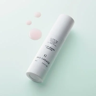 淡いミントグリーンの背景に、白いボトルに入ったスキンケア製品「AND CREAGE STEM AWAKEN BOOSTER」が置かれています。製品の隣にはピンク色の液体が数滴垂れており、シンプルで清潔感のあるイメージです。