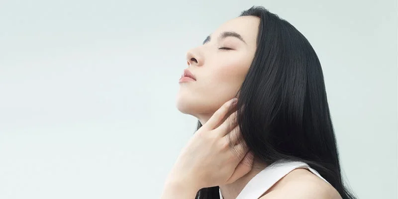 目を閉じ、上を向いている女性の横顔のポートレート。長い黒髪で、片手を首元に添え、穏やかな表情を見せており、透明感のある肌が印象的です。