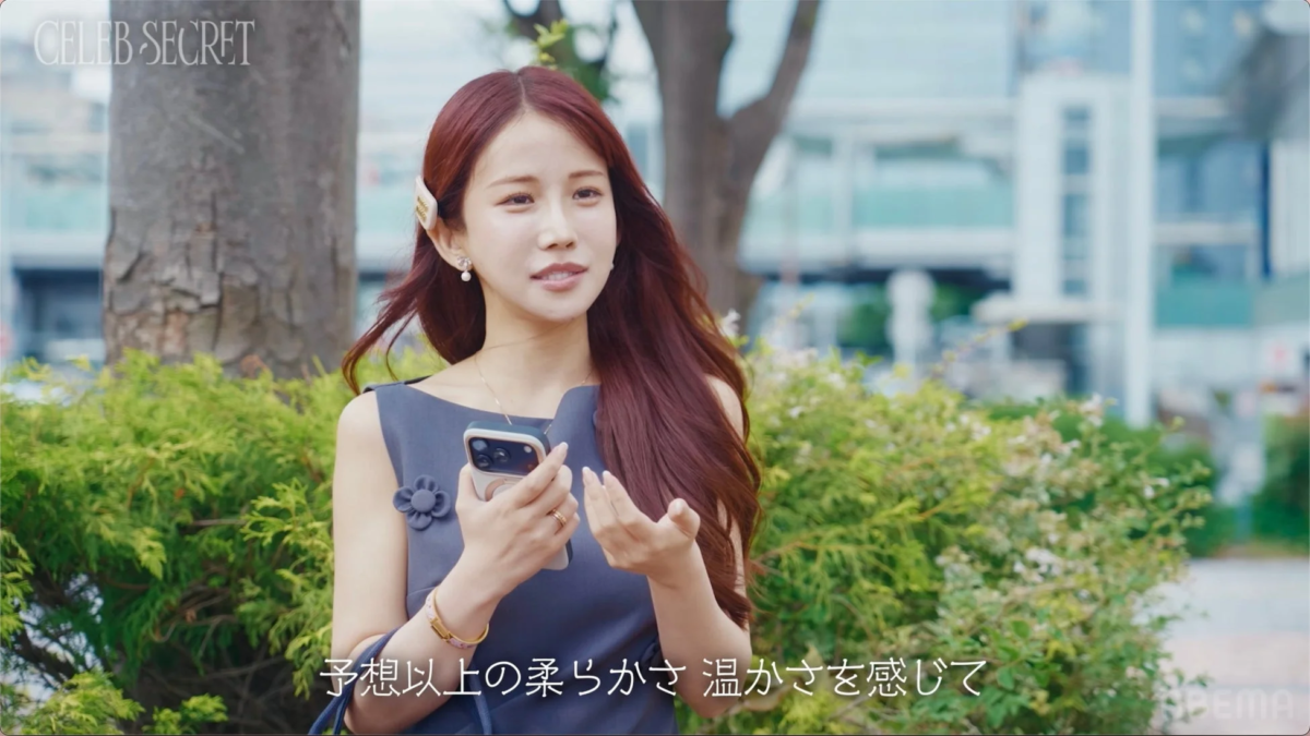 屋外でスマートフォンを手に、穏やかな表情で話す女性のポートレート。背景には緑豊かな植え込みと街並みが広がる。字幕には「予想以上の柔らかさ 温かさを感じて」と表示されており、何かについて語っている様子がうかがえる。