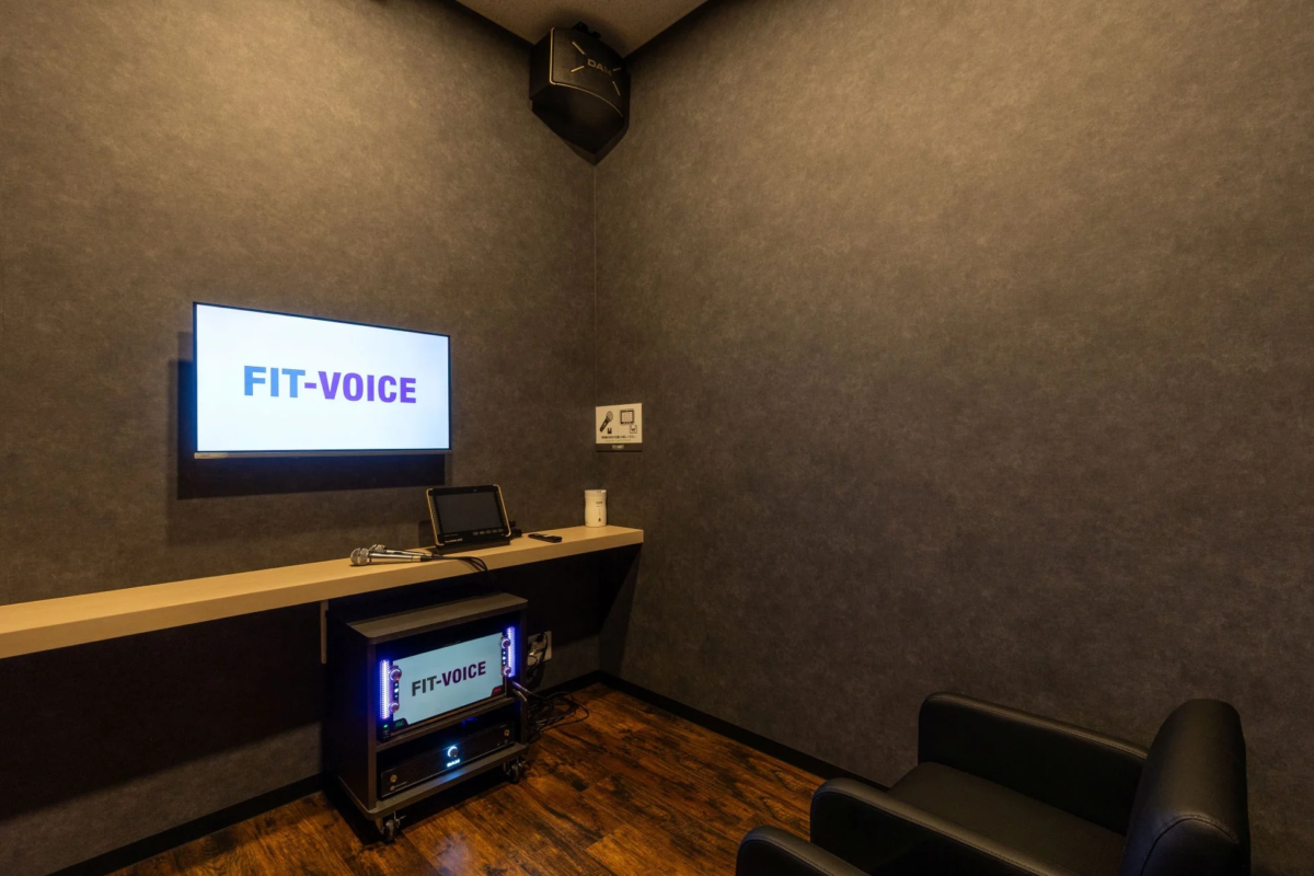 「FIT-VOICE」のロゴが表示されたスクリーンやマイク、音響機器が設置されたカラオケルーム