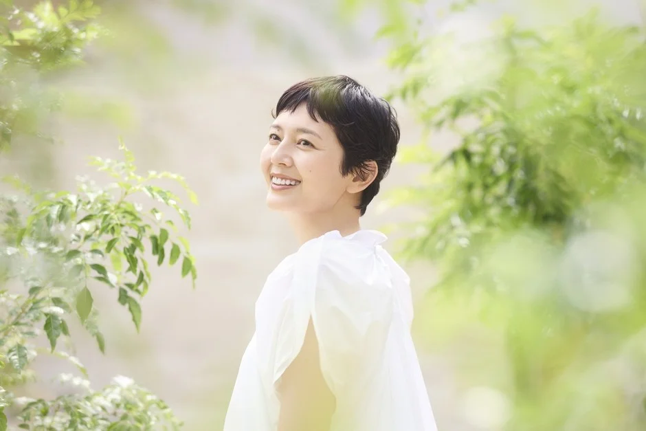A portrait of a short-haired woman in a white blouse looking up with a soft smile in bright sunlight. The green in the background gives a fresh impression.