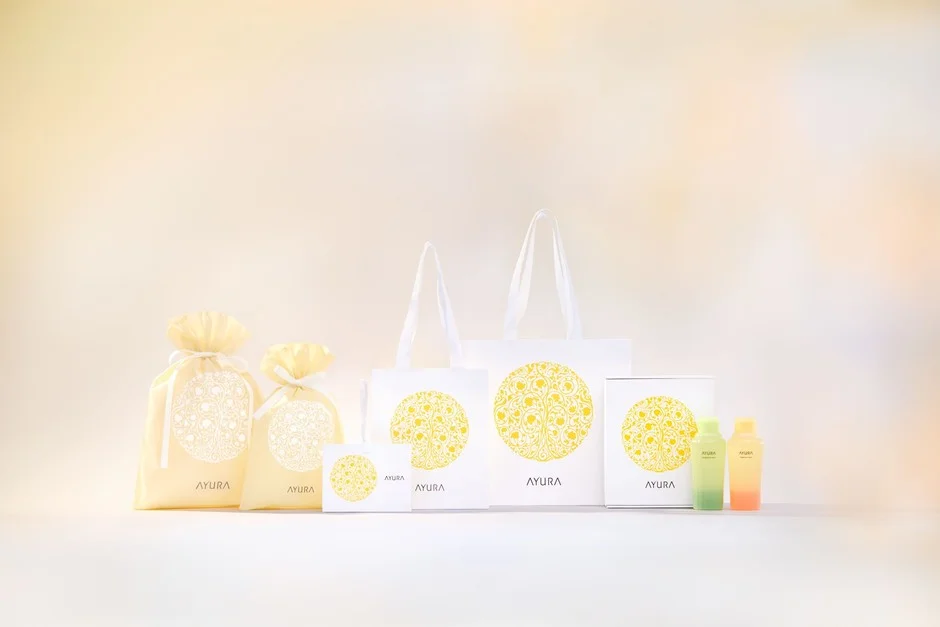 An image showing AYURA cosmetics and gift set packages, a tote bag, a drawstring pouch and a small bottle arranged together. The unified design in white and yellow gives a clean, elegant feel.