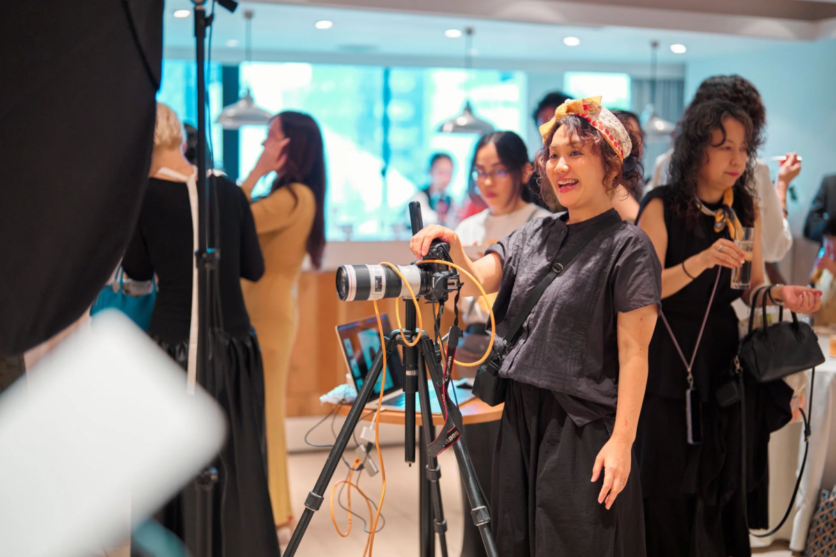 ヘッドスカーフを巻いた女性が、三脚に据えられたプロ用カメラの横で笑顔を見せています